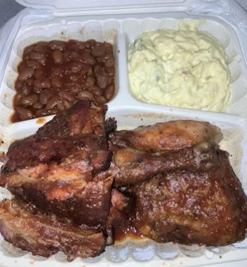 A takeout container with BBQ ribs, chicken, baked beans, and potato salad.