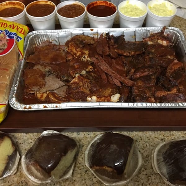 A large catering tray of barbecue ribs and brisket, with sides of beans, potato salad, bread, and cake.