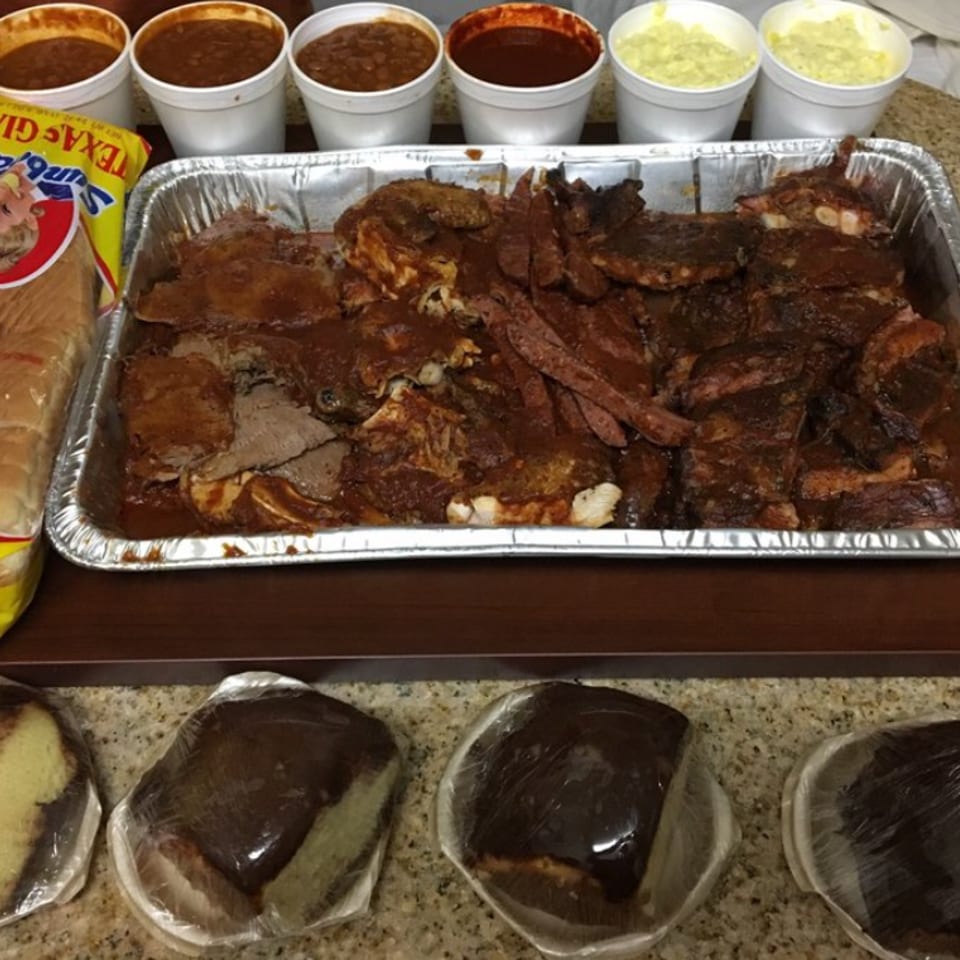 A large catering tray of barbecue ribs and brisket, with sides of beans, potato salad, bread, and cake.