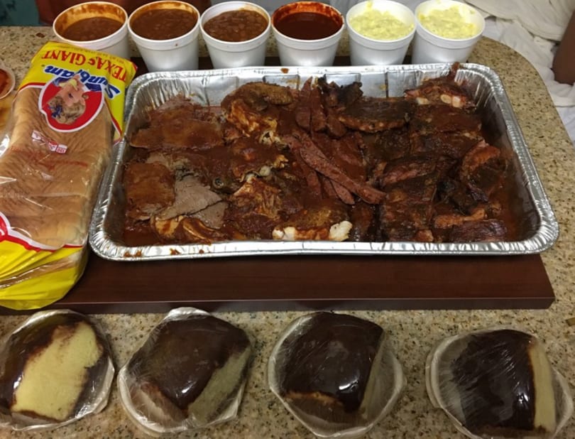 A large catering tray of barbecue ribs and brisket, with sides of beans, potato salad, bread, and cake.